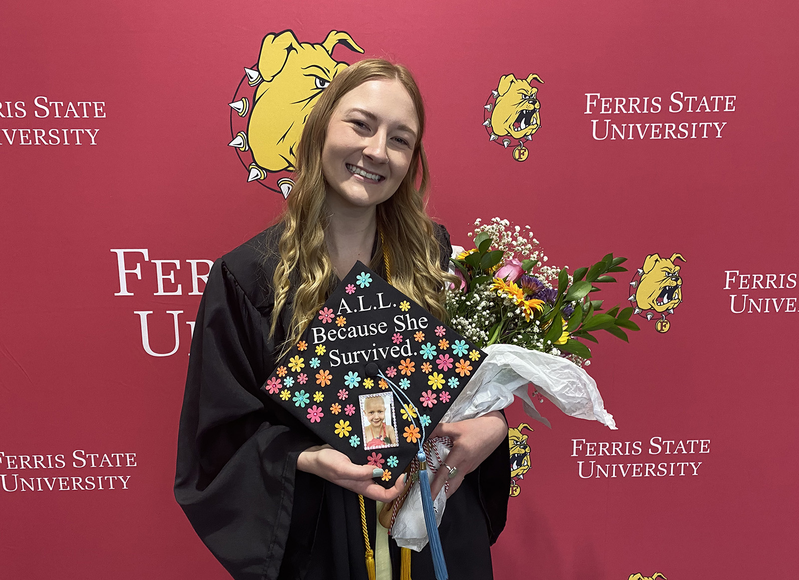 Recent Ferris State graduate Luci Archer holds up her graduation cap featuring the words "A.L.L. because she survived"